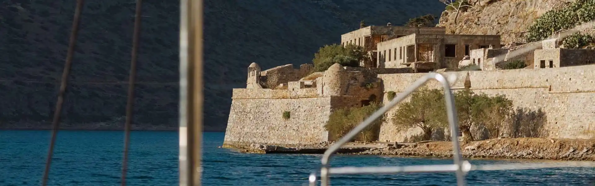 View of Spinalonga Island seen from the sea during a cruise departing from Agios Nikolaos near Elounda, Crete, with rocky shores and historic fortifications rising above the blue water.