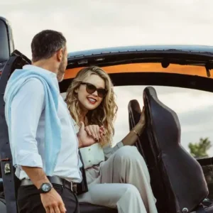 Woman smiling as she boards a sleek black helicopter, holding hands with a man while a pilot stands nearby.
