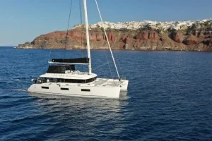 Luxury Lagoon 620 catamaran sailing in the Santorini Caldera with Oia village and white cliffside buildings in the background under a clear blue sky.