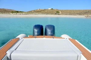 Rear view from a speedboat facing the turquoise waters of Rhenia Islet near Mykonos, with twin outboard engines and a quiet shoreline ahead.