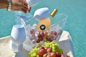 A chilled bottle of rosé wine with grapes and glasses is served on a speedboat above the clear turquoise waters of Mykonos.